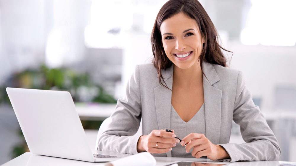 Kvinna i ljusgråa affärskläder sitter vid ett bord med sin laptop bredvid sig och ler in mot kameran