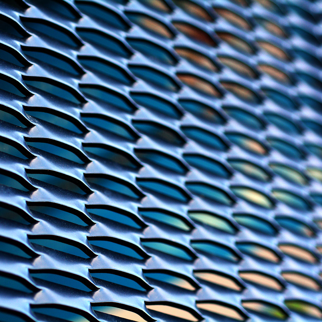 Närbild av ett perforerat metallnät med upprepade geometriska öppningar. Metallytan reflekterar blå, gröna och varma toner och skapar ett abstrakt industriellt mönster.