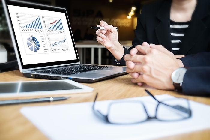En person visar diagram och statistik på en bärbar dator under en presentation, glasögon ligger på bordet framför.