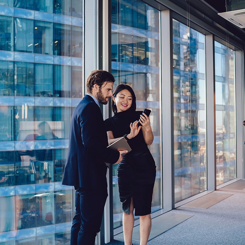Eine Frau und ein Mann stehen in einem modernen Bürogebäude und schauen gemeinsam auf ein Smartphone.