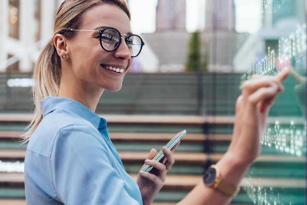 Femme debout devant un kiosque numérique interactif, smartphone à la main.