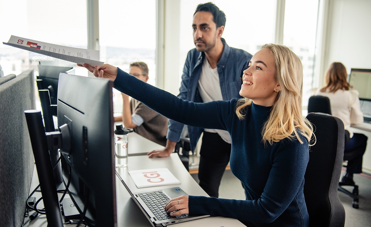 To kolleger peker på skjerm og samarbeider ved arbeidsstasjon. To kolleger peker på skjerm og samarbeider ved arbeidsstasjon.