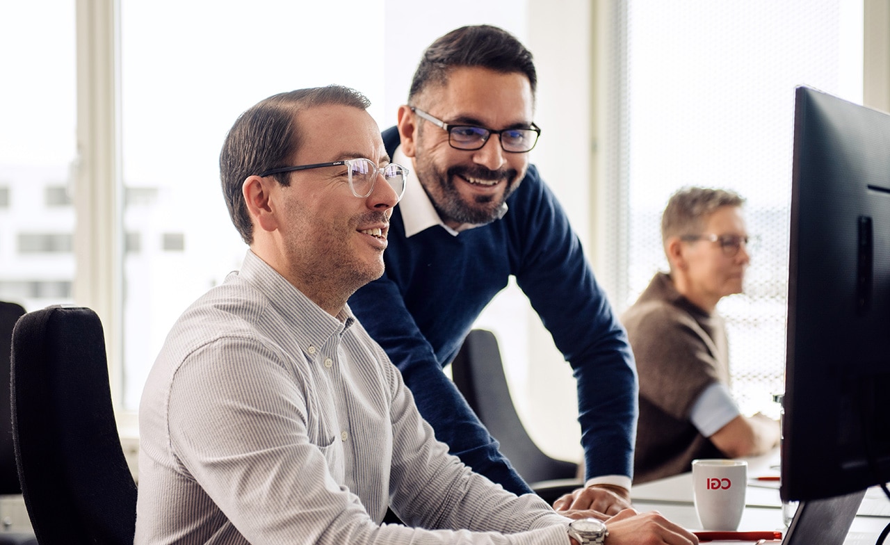 To personer smiler foran dataskjerm, tredje jobber i bakgrunnen med CGI-kopp på bordet.