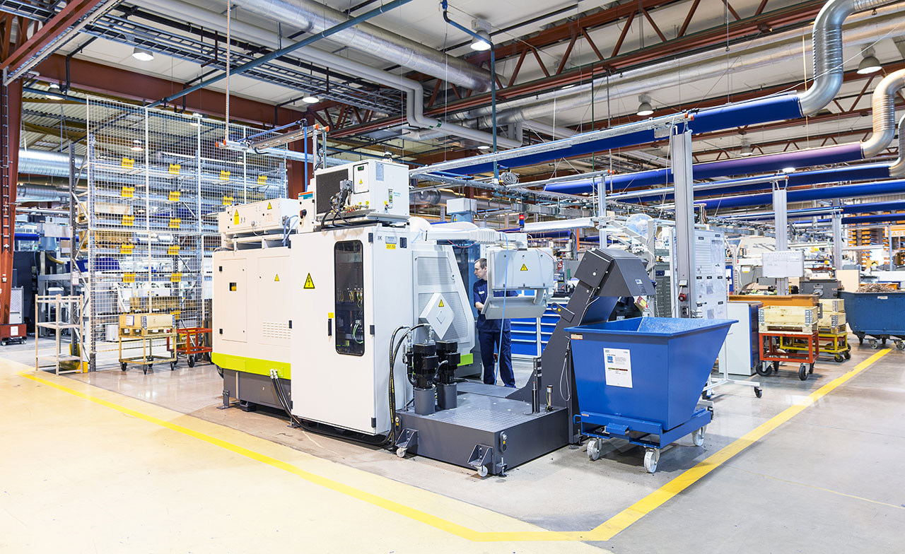 Interiör av en modern industrifabrik med stora produktionsmaskiner, teknisk utrustning och rörsystem. Produktionsområdet är ljust och organiserat med markerade säkerhetszoner på golvet.
