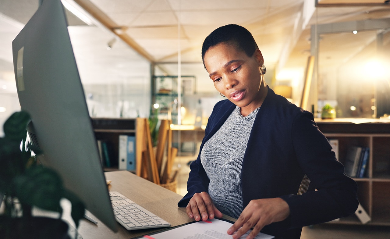 Person ser på dokument ved skrivebord med stasjonær PC.