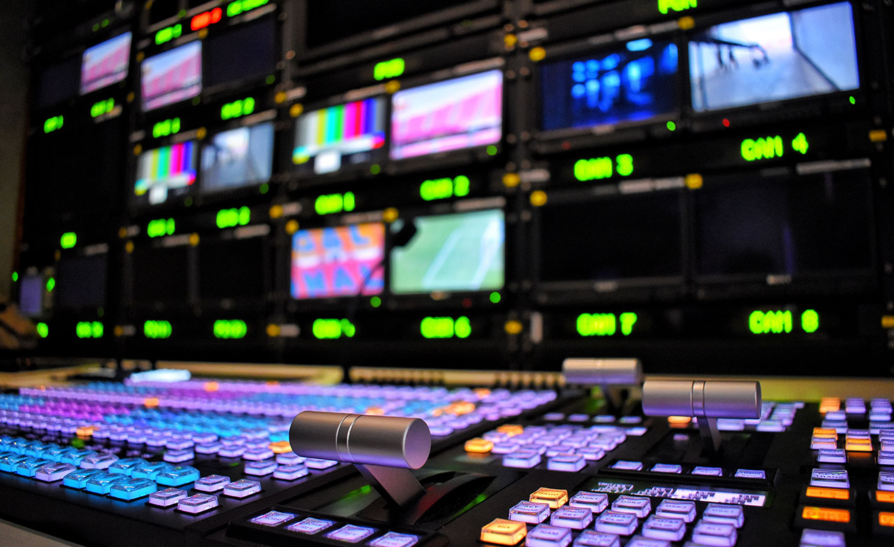 Kontrollrum för tv- eller videoproduktion med en mixerpanel i förgrunden och flera skärmar som visar olika videokällor i bakgrunden. Färgglada knappar och teknisk utrustning används för att styra sändning och produktion.