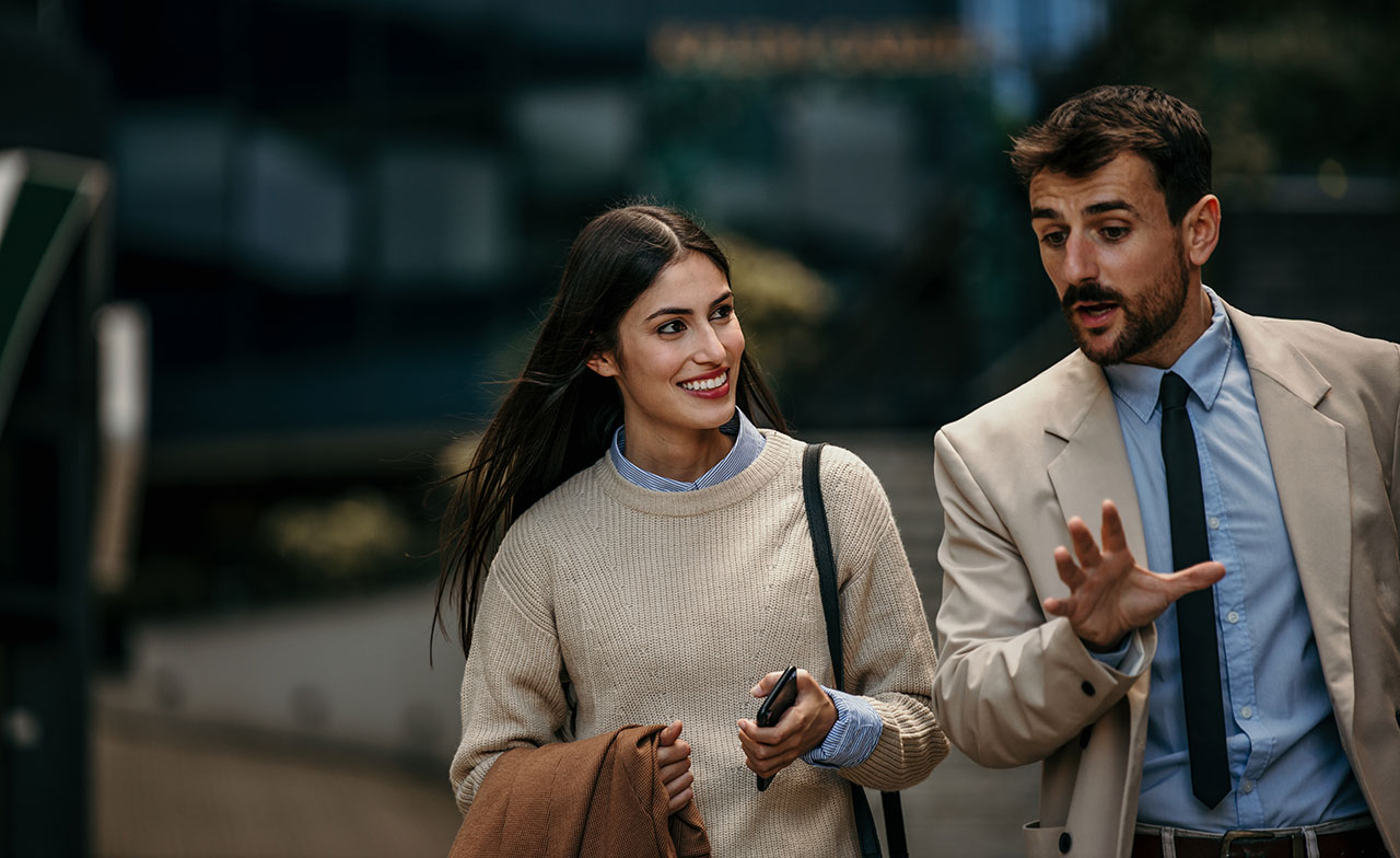 business_colleagues_talking_outdoors_city_walk_736565038 En man och en kvinna i affärskläder promenerar tillsammans utomhus i en stadsmiljö. Kvinnan håller i sin kappa och ler medan mannen pratar engagerat. Båda ser ut att vara i ett samtal på väg till eller från arbete.