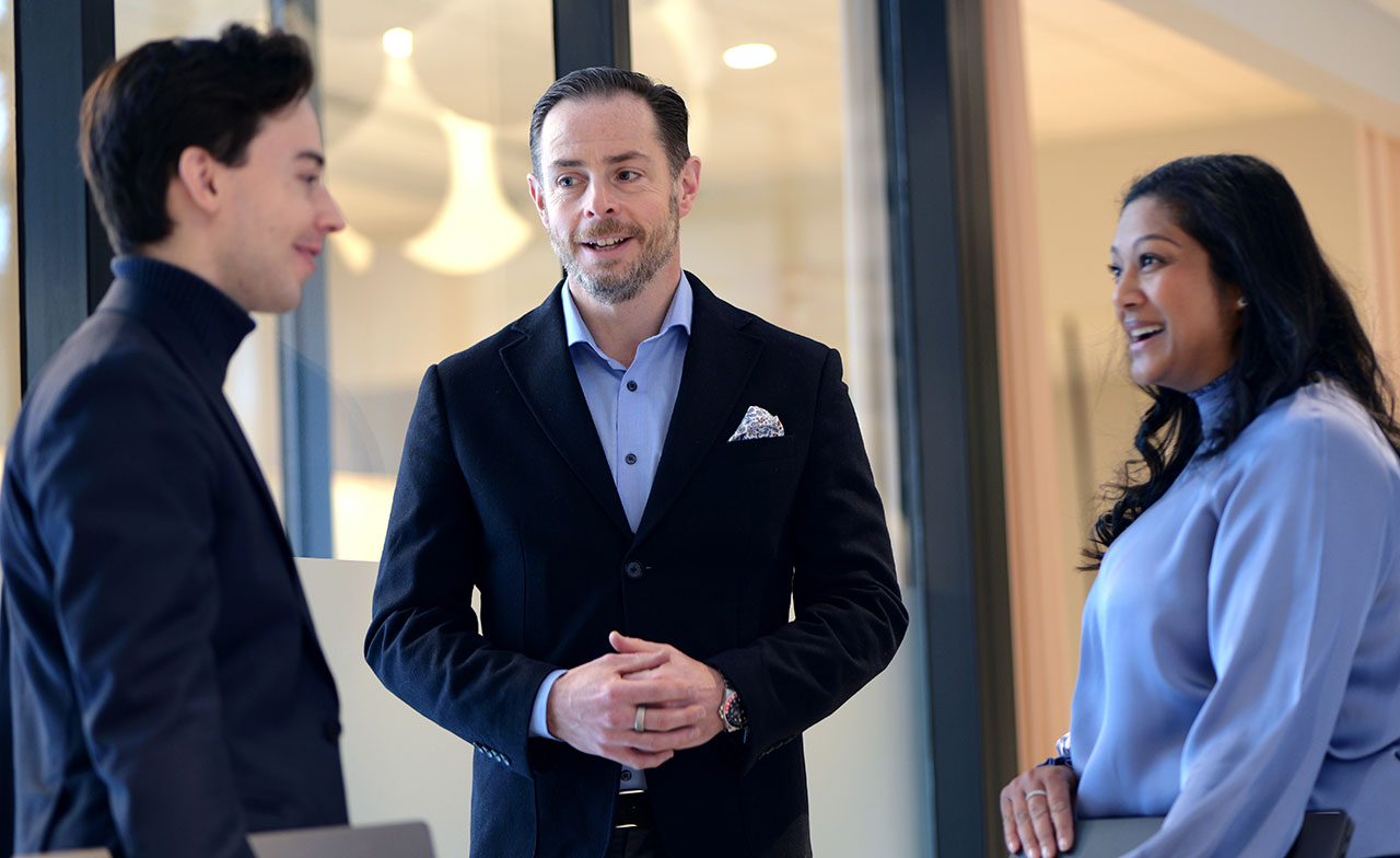 Tre personer i affärskläder står och samtalar i en modern kontorsmiljö. De ler och verkar ha en avslappnad och professionell dialog.