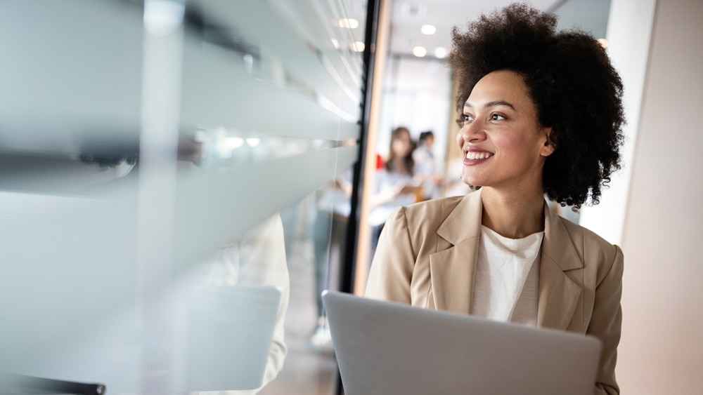 A businesswoman smiling while standing with a laptop in an office window