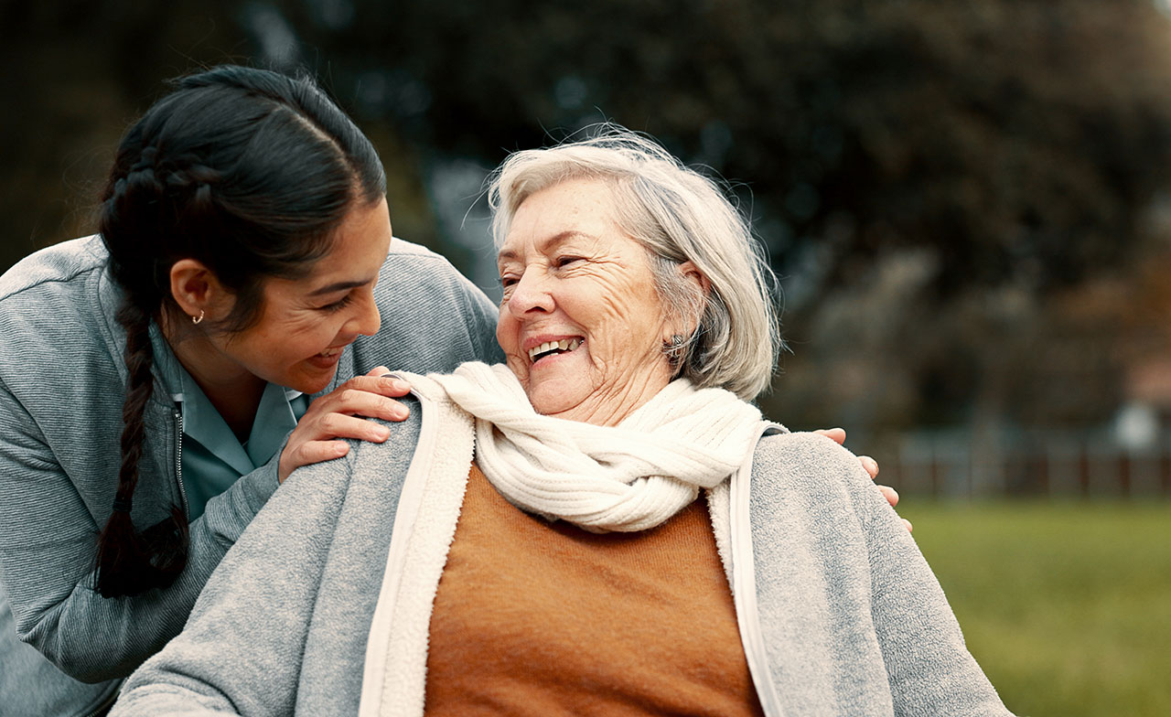 caregiver_smiling_with_elderly_woman_outdoors En yngre kvinna, troligen en vårdgivare, ler varmt mot en äldre kvinna i rullstol. De håller om varandra och skrattar tillsammans utomhus i en grön parkmiljö.