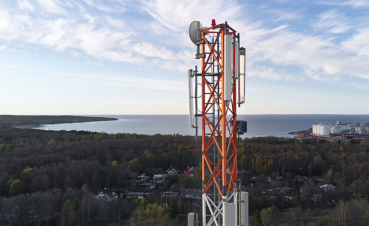 En mobilmast med antenner står i ett kustnära landskap med skog och vatten i bakgrunden. Bilden visar telekommunikationsinfrastruktur i en naturnära miljö.