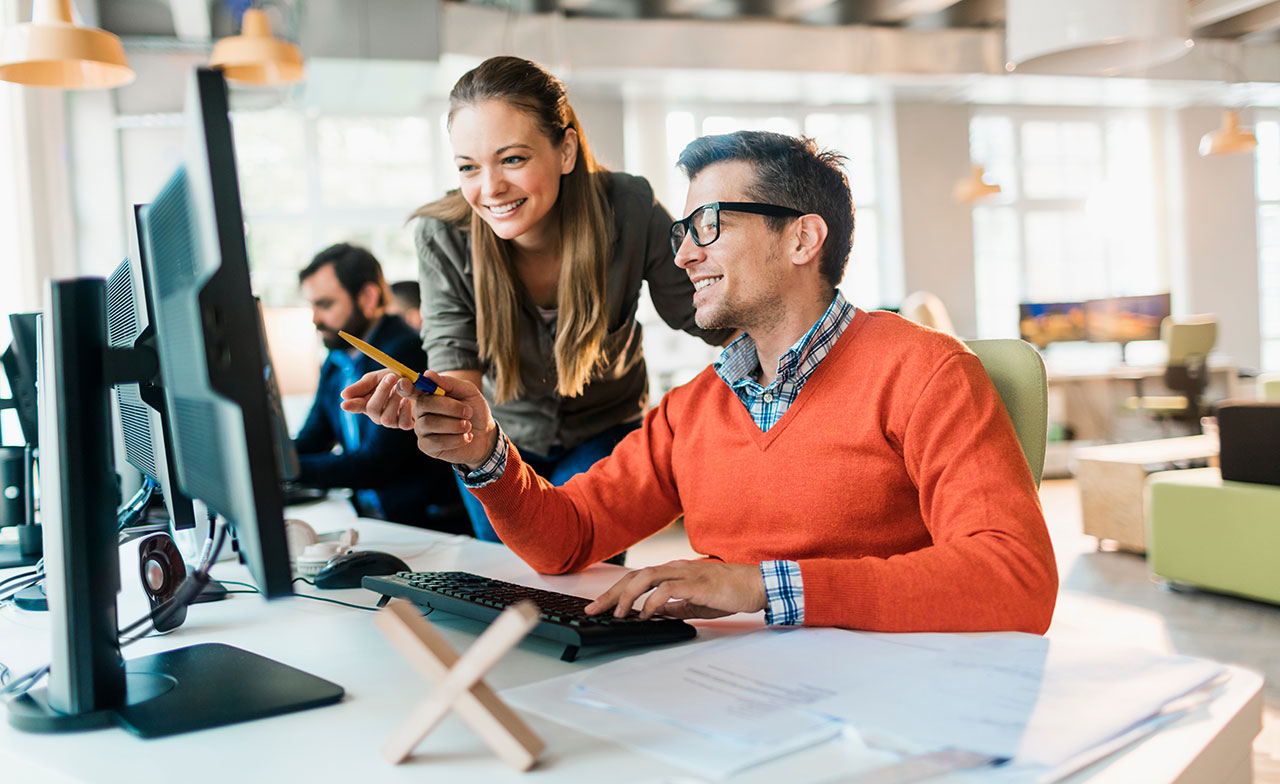 Två kollegor samarbetar vid en dator i ett modernt kontor och diskuterar arbetet framför skärmen.