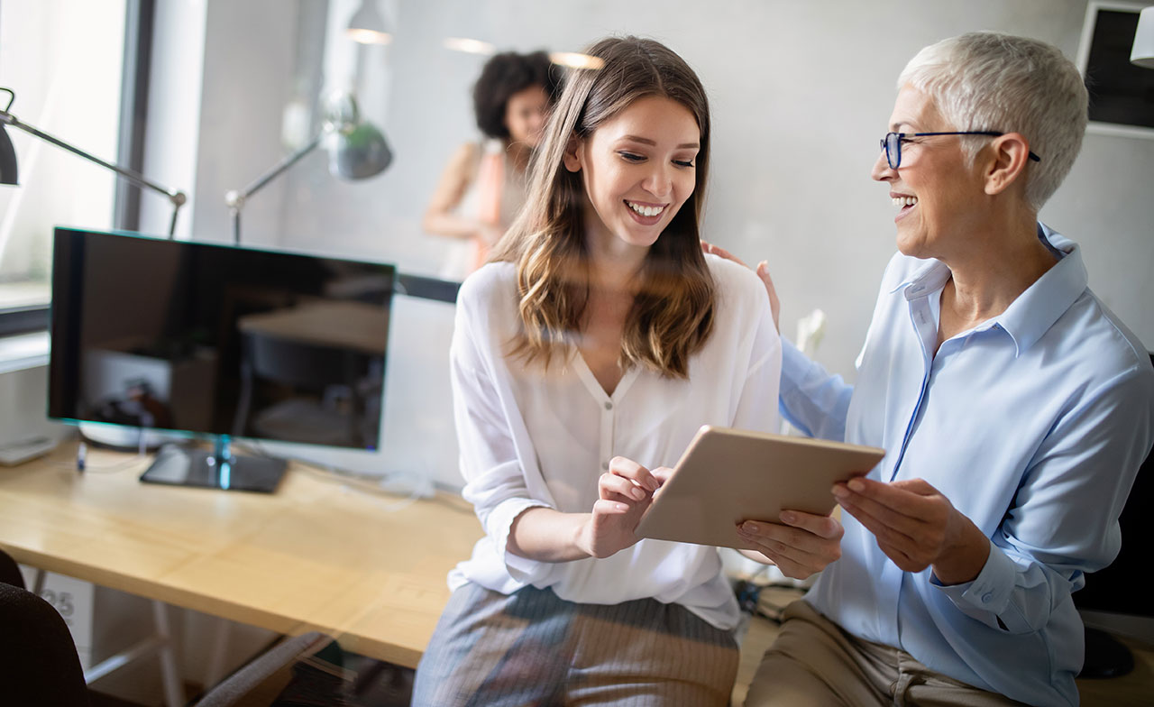 Två kvinnor i olika åldrar samarbetar leende framför en surfplatta på ett modernt kontor. En sitter på skrivbordet, den andra står bredvid, och i bakgrunden syns ytterligare en kollega. Stämningen är positiv och stödjande.