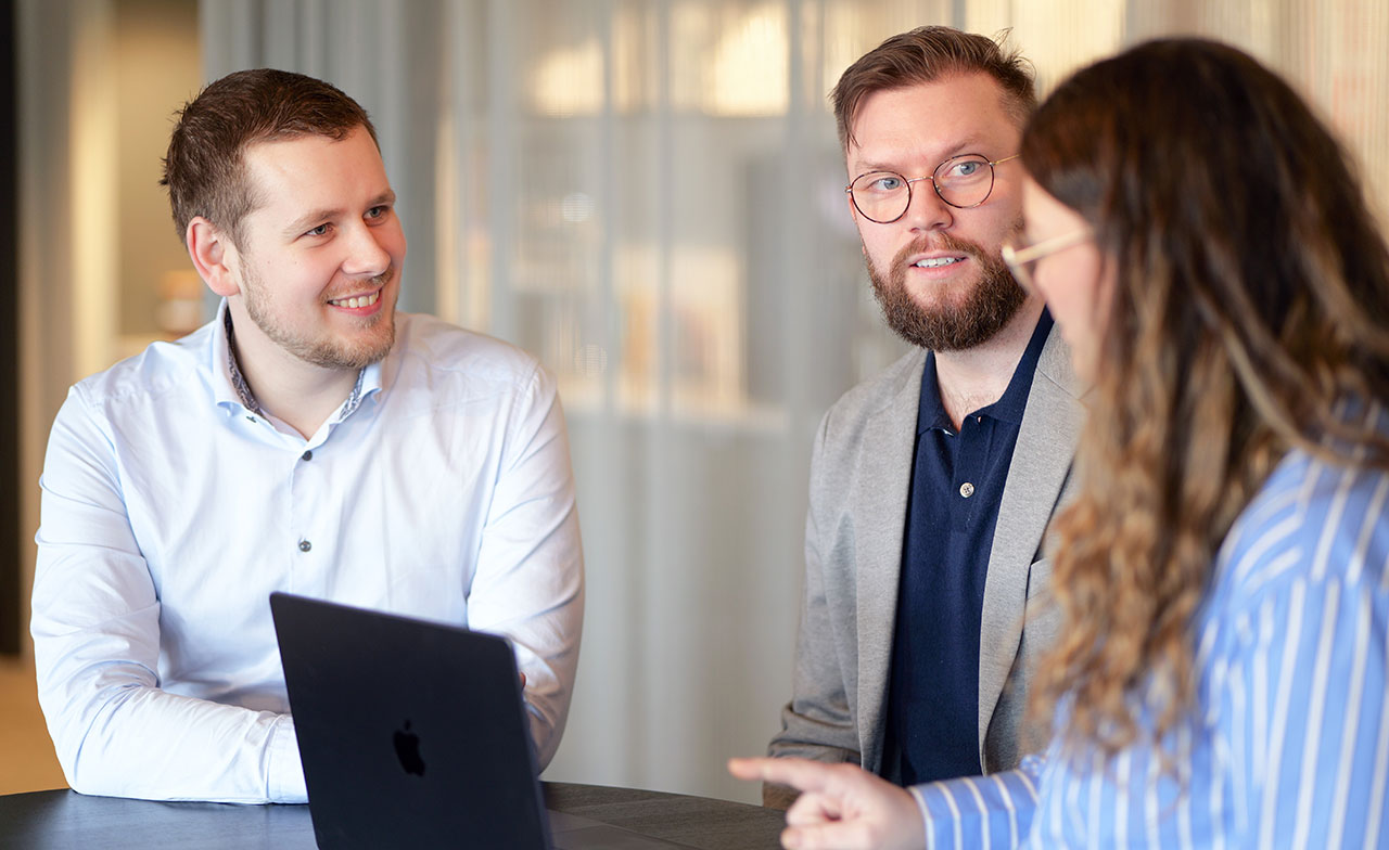 Tre kollegor sitter tillsammans vid ett bord och diskuterar kring en laptop. De ler och verkar engagerade i samtalet i en modern kontorsmiljö.