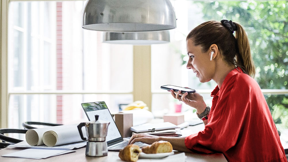 Kvinna i röd blus sitter vid sitt frukostbord och pratar i mobiltelefon samtidigt som hon tittar på sin laptop