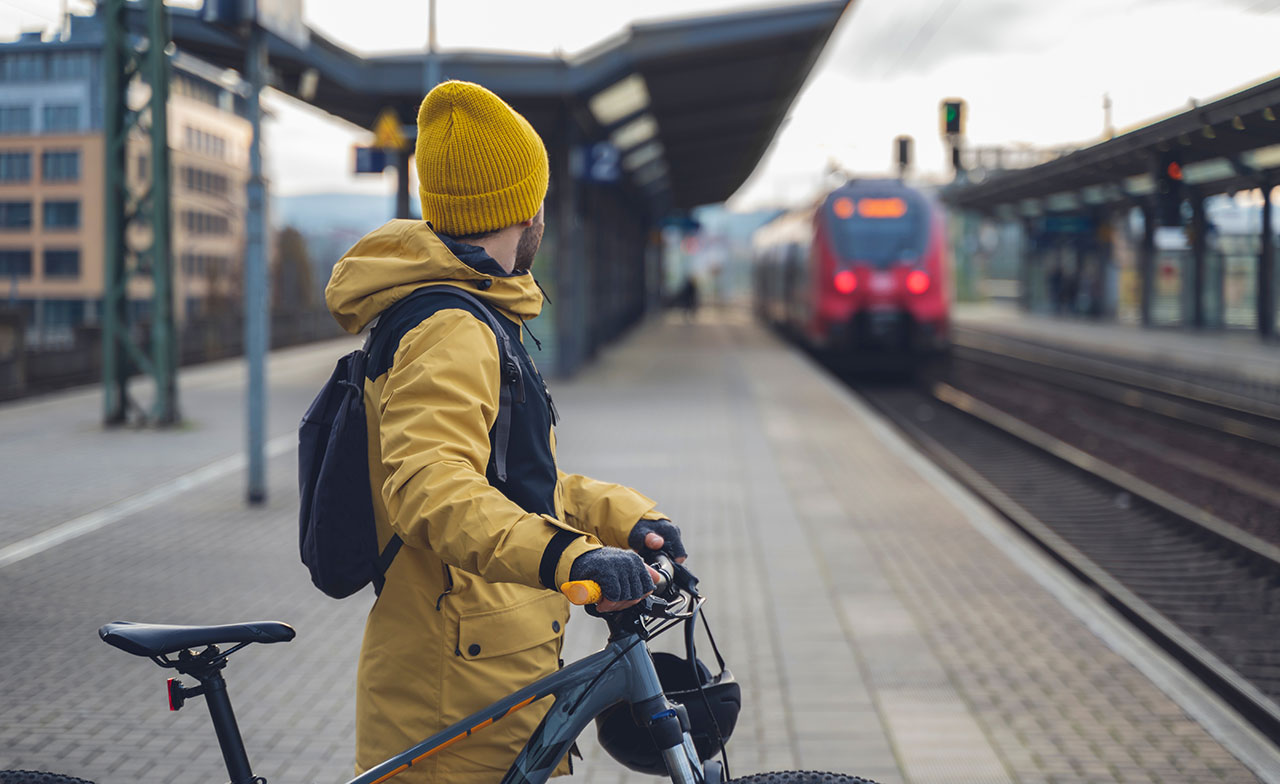 commuter_with_bike_waiting_train_station_394754304 En person i gul jacka och gul mössa står på en tågperrong med en cykel, och tittar mot ett inkommande rött tåg. Personen bär ryggsäck och verkar vara på väg att resa vidare med kollektivtrafik.