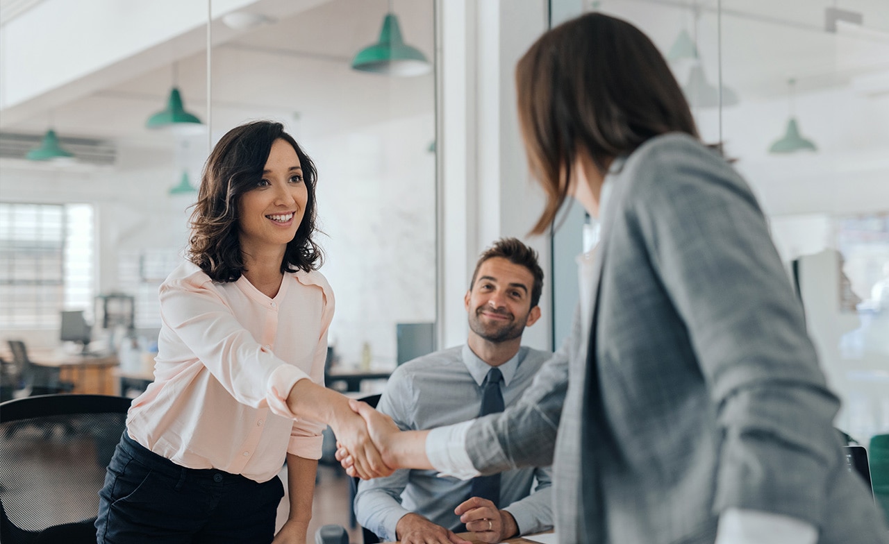 To personer håndhilser i kontorlokale, tredje person ser på og smiler