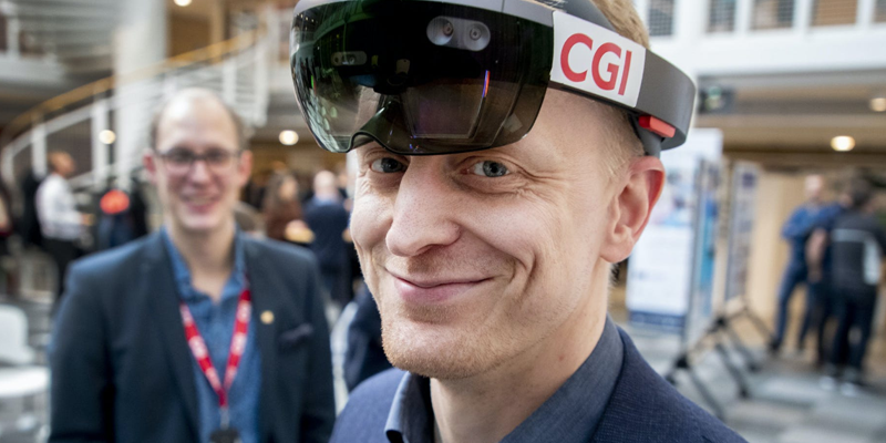 CGI-medarbetare ler in mot kameran med VR-glasögon uppdragna i pannan. I bakgrunden syns en annan kollega som också står i montern på en mässa