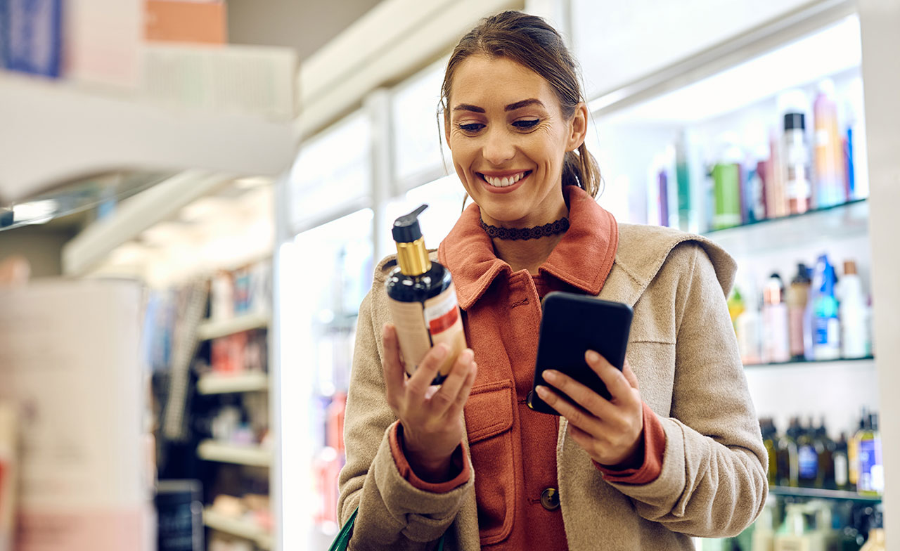 En kund i en butik håller en produkt i ena handen och använder en smartphone i den andra. Hon ler medan hon granskar produktinformation på mobilen framför butikshyllorna.