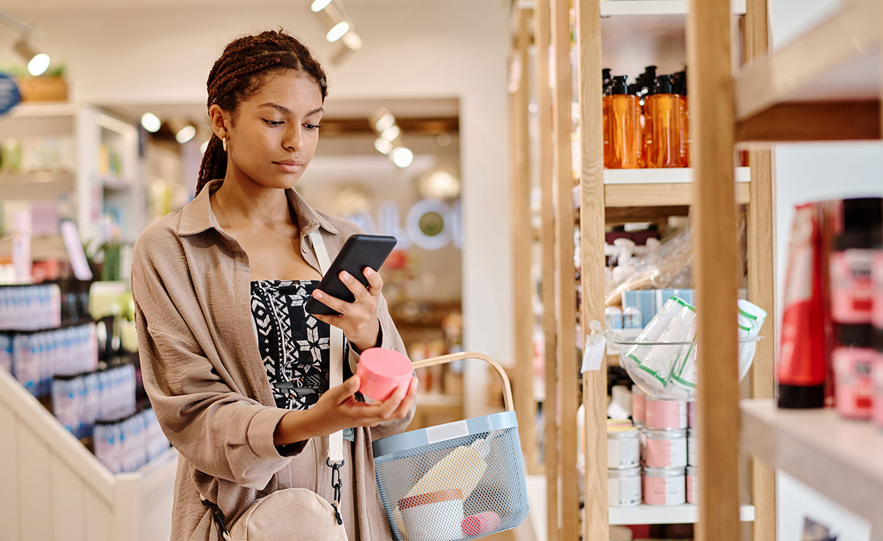 En kund står i en butik och använder sin mobiltelefon samtidigt som hon håller en produkt i handen. Hyllor med olika varor syns runt omkring henne.