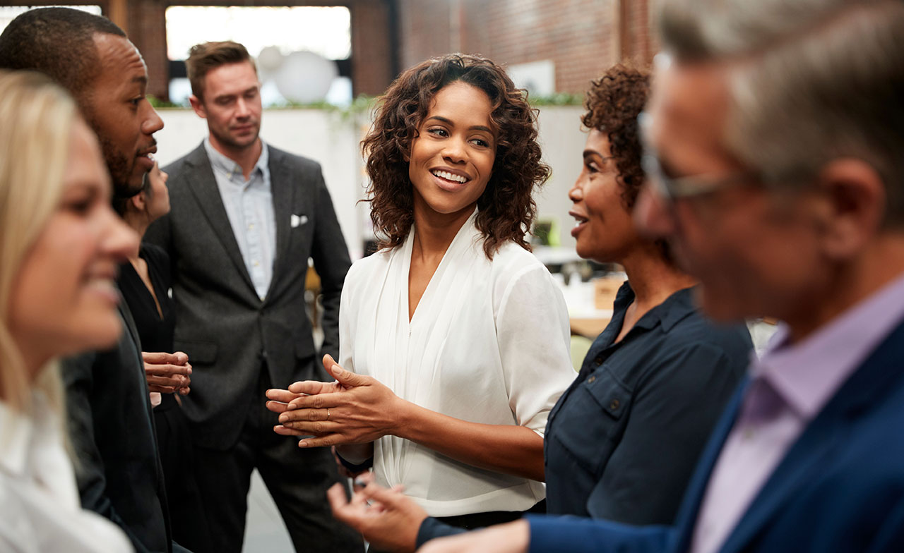 diverse_professionals_networking_event_smiling_257397505 En grupp yrkespersoner samtalar och skrattar tillsammans vid ett nätverksevent i en modern inomhusmiljö. Fokus ligger på en leende kvinna i vit skjorta i mitten, omgiven av kollegor i affärskläder.