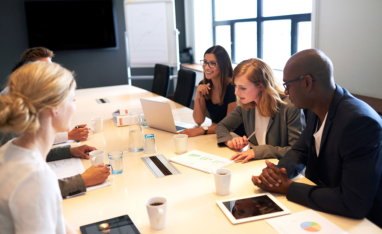 En grupp kollegor sitter runt ett konferensbord och samarbetar. De diskuterar dokument och arbetar med laptops och surfplattor i ett modernt mötesrum.