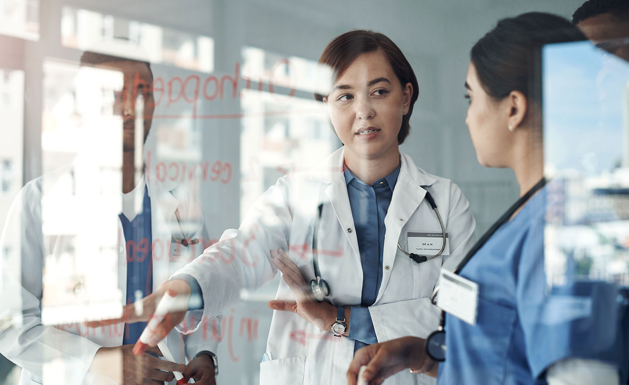 En grupp läkare diskuterar en behandlingsplan vid en genomskinlig glasvägg med anteckningar, där en läkare pekar och förklarar för sina kollegor.