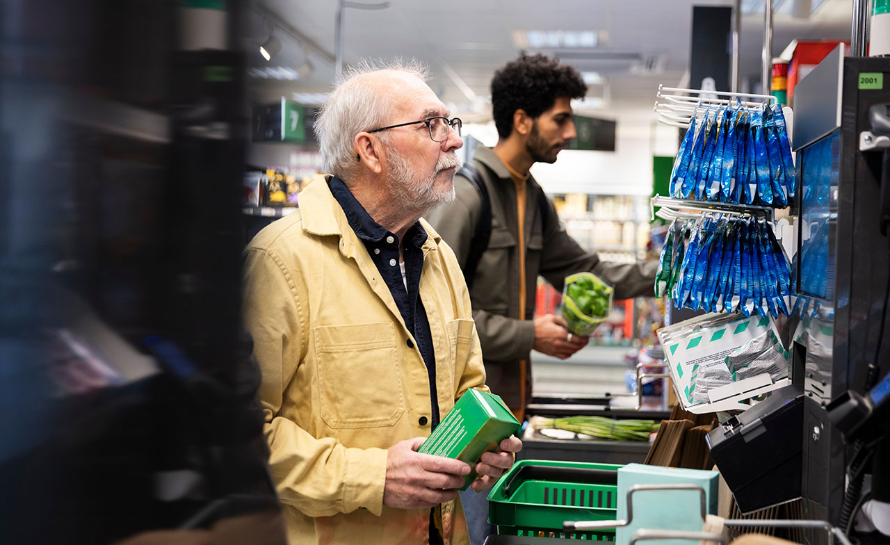 En äldre man står vid en självutcheckningskassa i en matbutik och håller i varor. I bakgrunden syns en yngre man med en grönsak i handen. Miljön är vardaglig och modern, med fokus på självbetjäning i handeln.