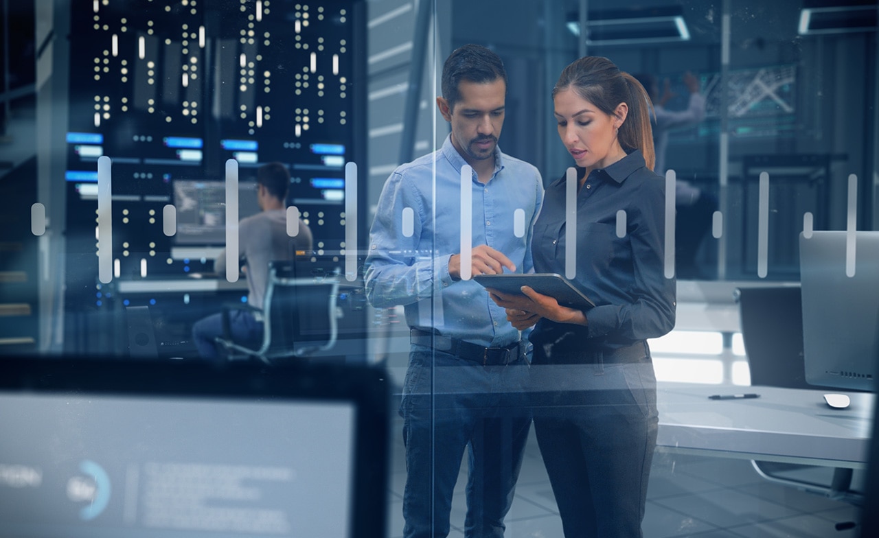 To personer ser på nettbrett i datasenter, omgitt av skjermer og servere.