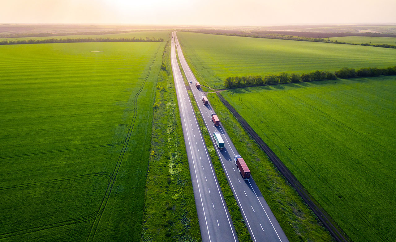 freight_trucks_highway_green_landscape_283383454 Flygfoto över lastbilar och andra fordon som kör på en lång motorväg genom ett vidsträckt grönt jordbrukslandskap. Bilden illustrerar vägtransport, logistik och godstrafik.