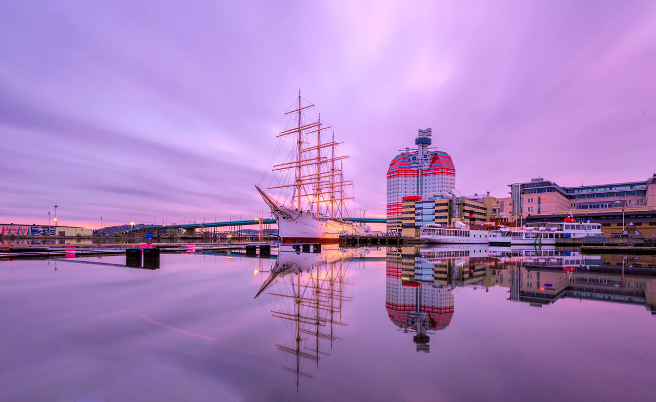 Vy över Göteborgs hamn i kvällsljus med segelfartyget Barken Viking och byggnaden Läppstiftet speglade i det stilla vattnet. Himlen är färgad i rosa och lila toner.