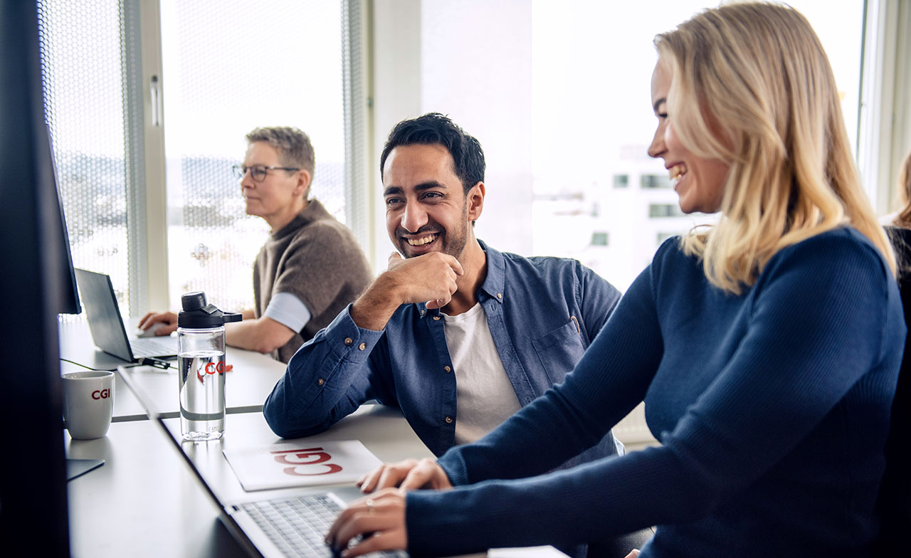 Tre kollegor arbetar vid sina datorer i ett ljust kontor. Två av dem, en man och en kvinna i förgrunden, samarbetar och skrattar tillsammans framför en datorskärm. Stämningen är positiv och fokuserad.