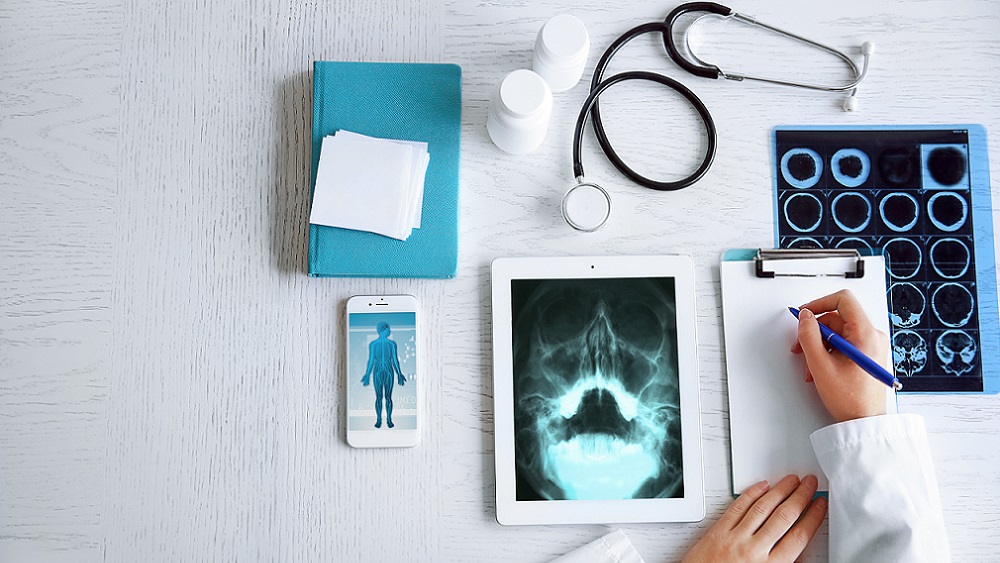 Läkare som tar noteringar vid sitt arbetsbord omgiven av surfplatta, mobil, medicinburkar, stetoskop och röntgenplåtar