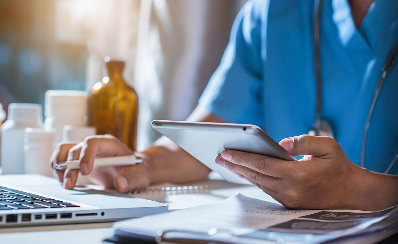 healthcare_professional_using_tablet_and_laptop_294289665 Närbild av vårdpersonal i blå arbetskläder som använder en surfplatta vid ett skrivbord med laptop, anteckningar och medicinsk utrustning. Bilden illustrerar digitalt arbete och informationshantering inom vården.