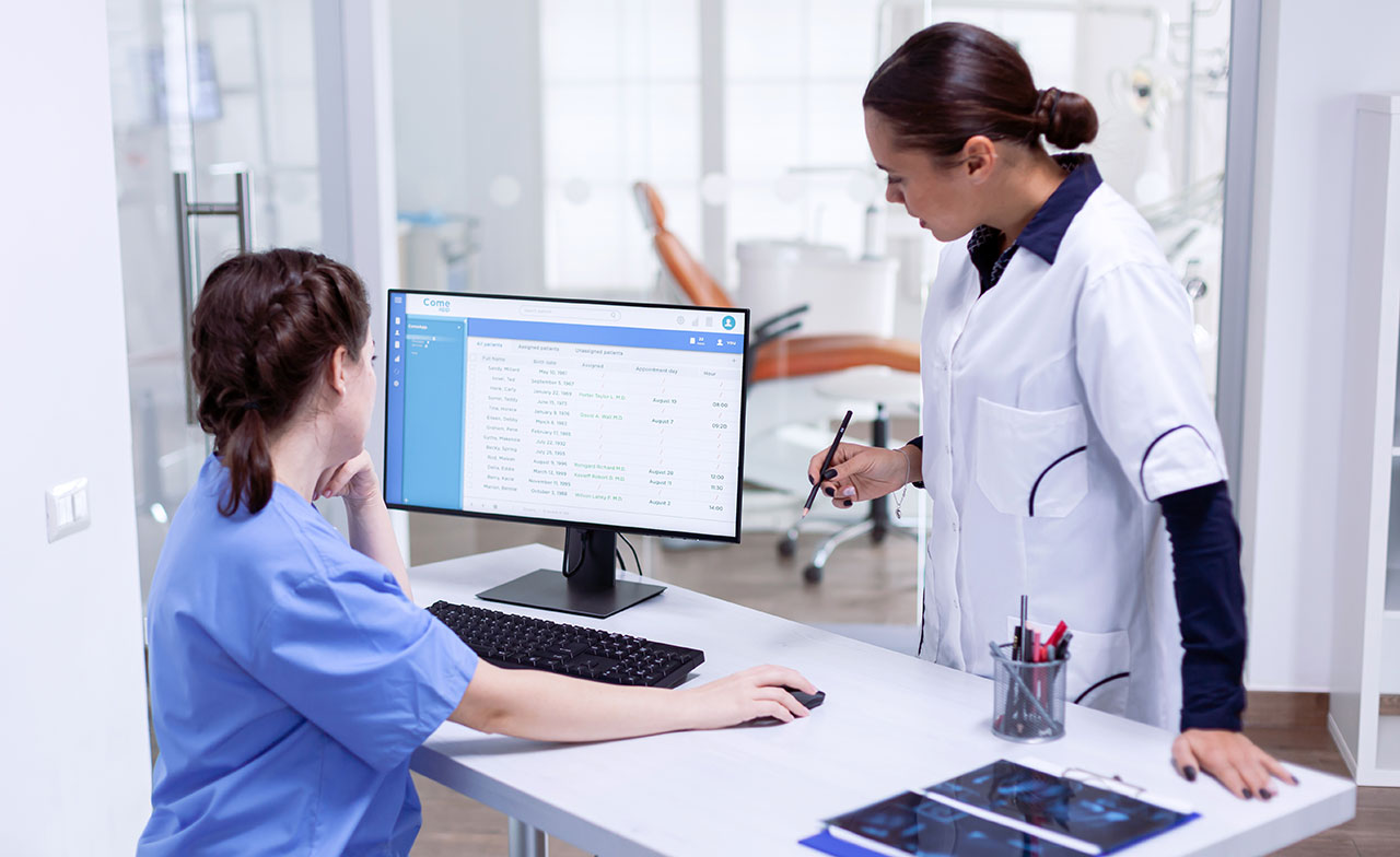 Två vårdpersonal i arbetskläder står vid ett skrivbord på en klinik och granskar patientinformation på en datorskärm. En sitter vid datorn och den andra pekar på skärmen med en penna. På bordet ligger även röntgenbilder.