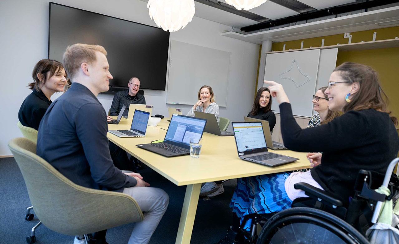 Sju kollegor sitter runt ett konferensbord i ett modernt kontor och deltar i ett möte. Flera laptops står på bordet och deltagarna ler och samtalar. En av kollegorna sitter i rullstol och gestikulerar under diskussionen.