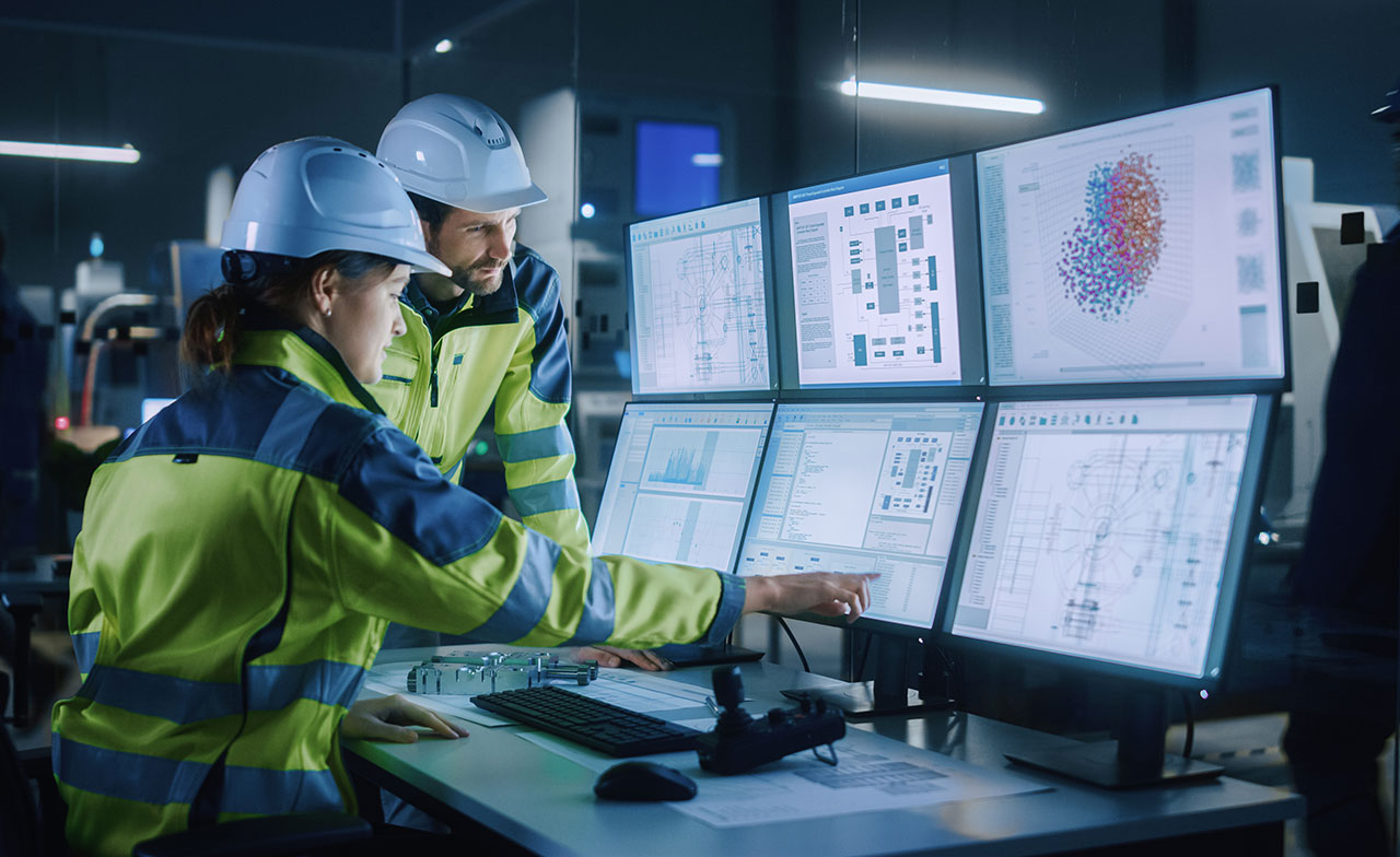 Två industriarbetare i hjälm och reflexjackor analyserar data på flera datorskärmar i ett kontrollrum. Skärmarna visar diagram, systemöversikter och teknisk information i en modern industrimiljö.
