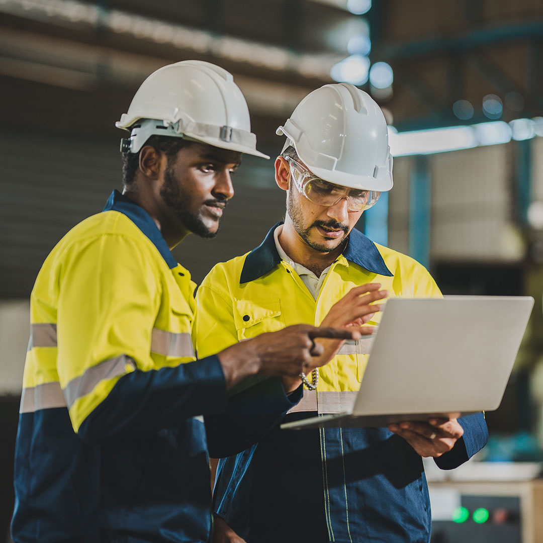 Två industriarbetare i skyddshjälmar och reflexkläder står tillsammans i en fabrik och granskar information på en laptop. Den ena pekar på skärmen medan de diskuterar arbetet.