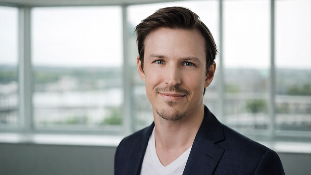 Porträtt av en man i kavaj och vit t-shirt som står framför stora fönster i en modern kontorsmiljö. Han ser in i kameran och ler svagt.