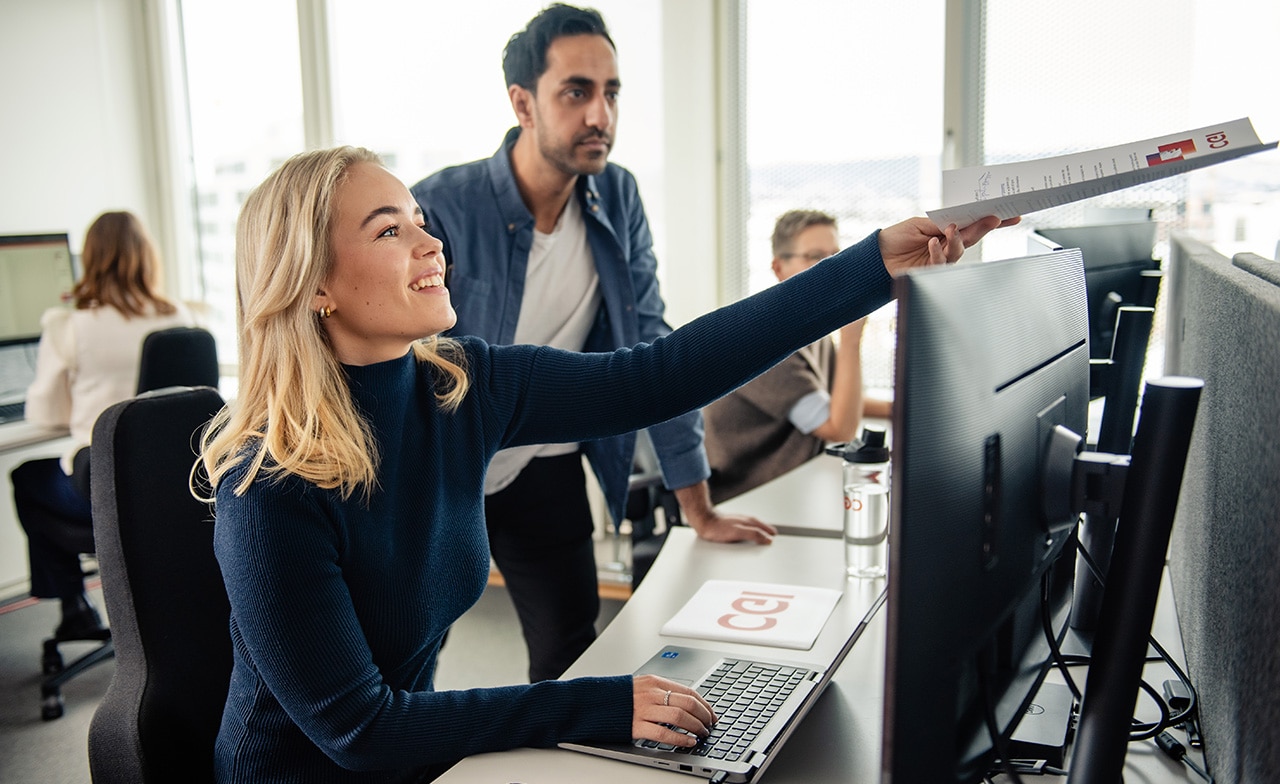 To kolleger peker på skjerm og samarbeider ved arbeidsstasjon.