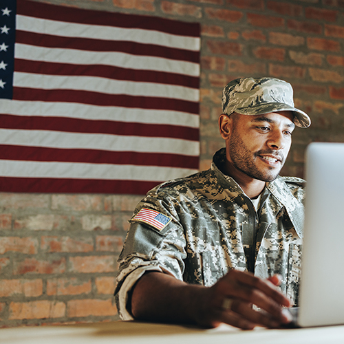 soldier at a laptop with American flag on the wall behind him