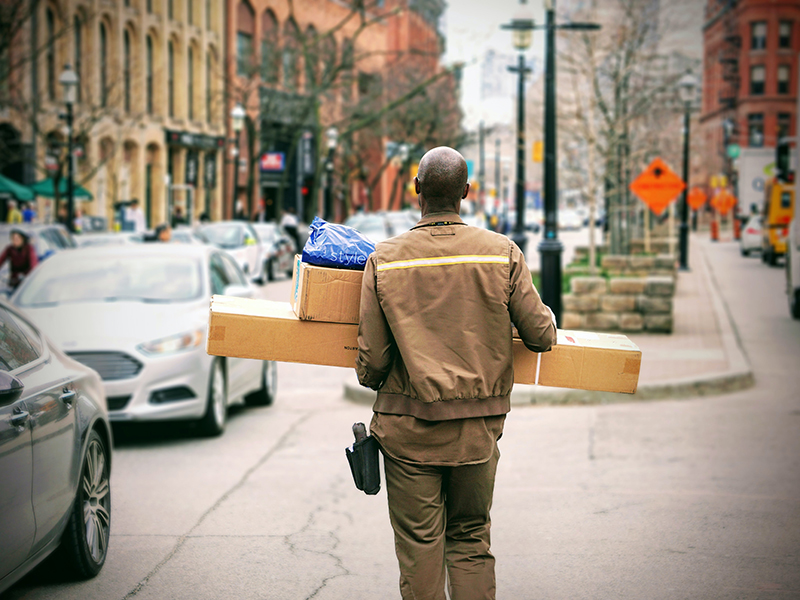 En man i bruna budbärarkläder ses gående bakifrån med famnen full av paket, han går på en gata i en storstad där både bilar och gaturarbete pågår 