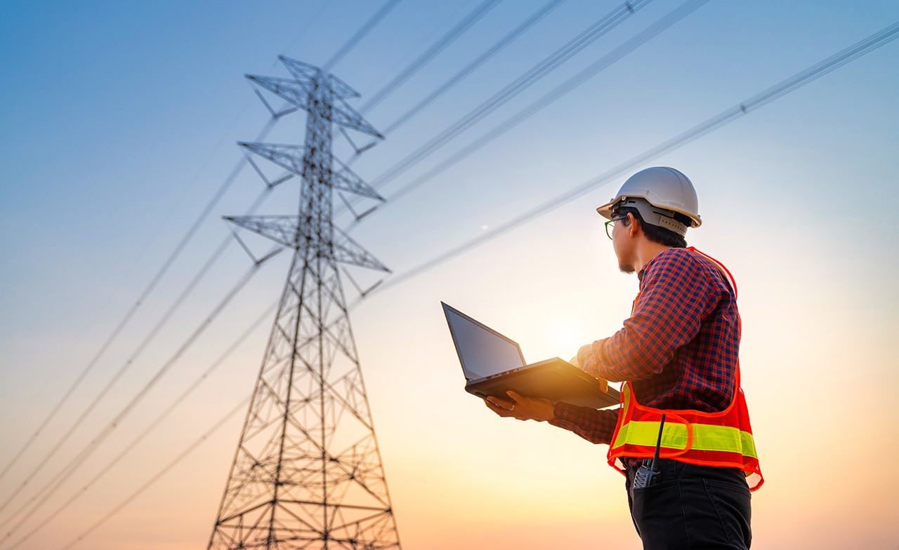 Arbeider med hjelm bruker laptop foran høyspentmast ved solnedgang. Arbeider med hjelm bruker laptop foran høyspentmast ved solnedgang.