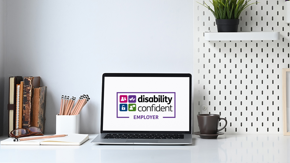 Disability confident logo displayed on a laptop on a desk with stationary and mug