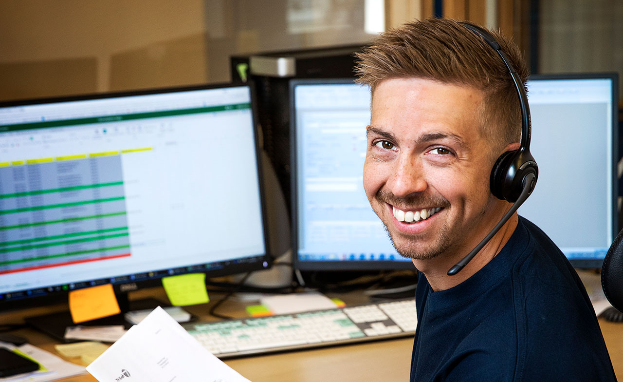 En leende person med headset sitter vid en dator med flera skärmar och tittar mot kameran. Bilden visar kundsupport eller IT-support i en kontorsmiljö.