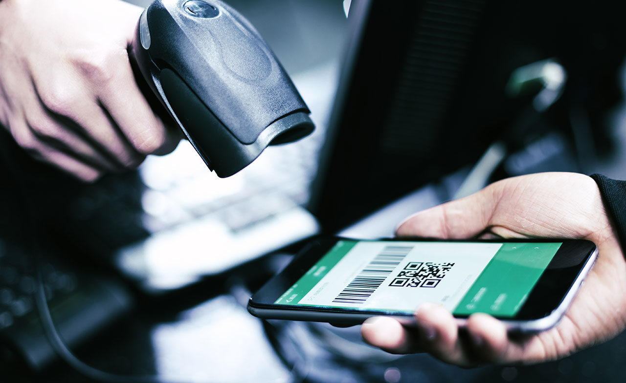 Närbild av en smartphone som visar en streckkod och QR-kod medan en handscanner riktas mot skärmen. Bilden illustrerar digital skanning och mobil identifiering i butiksmiljö.