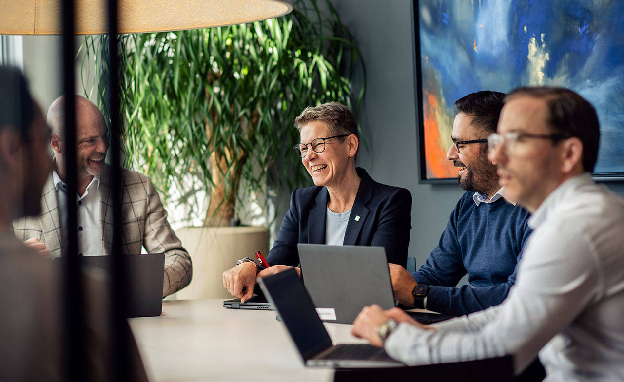 En grupp kollegor har ett möte runt ett bord i ett modernt kontor i Norge. Alla har bärbara datorer framför sig och samtalet sker i en avslappnad och positiv atmosfär. En stor grön växt och färgstark konst syns i bakgrunden.