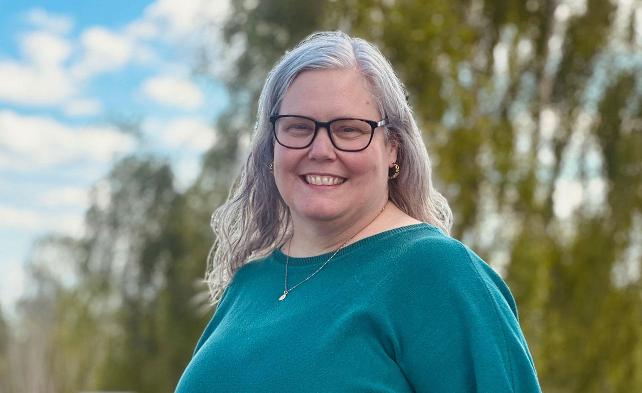 Porträtt av en leende kvinna med glasögon och långt grått hår som står utomhus. I bakgrunden syns träd och natur i mjukt fokus, vilket ger bilden en varm och välkomnande känsla.
