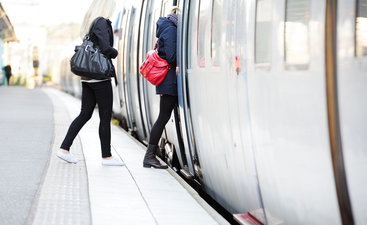 Två personer kliver på ett tåg vid en perrong. De bär väskor och är på väg in genom tågdörrarna, vilket visar vardagspendling med kollektivtrafik.