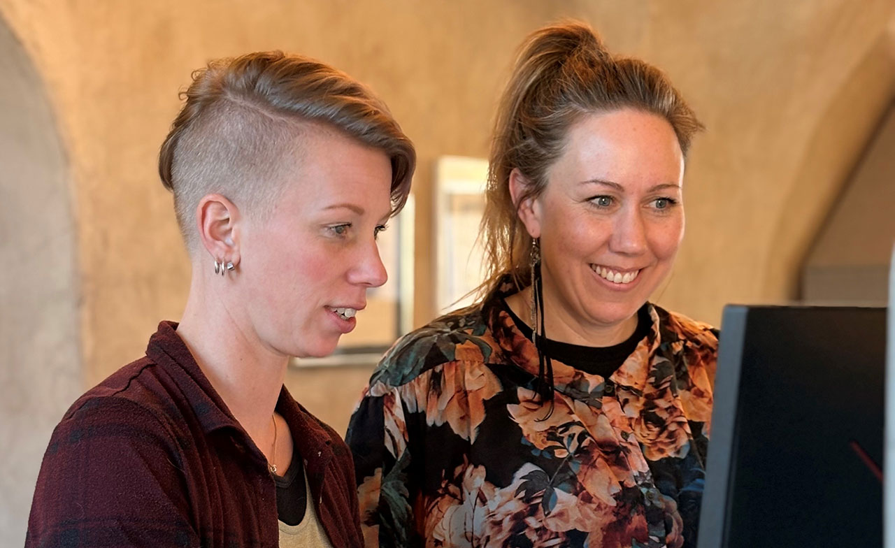 Två kvinnor står bredvid varandra och tittar på en datorskärm. Den ena pekar eller fokuserar koncentrerat medan den andra ler. De befinner sig i en varm och informell inomhusmiljö, vilket signalerar samarbete och gemensamt arbete.